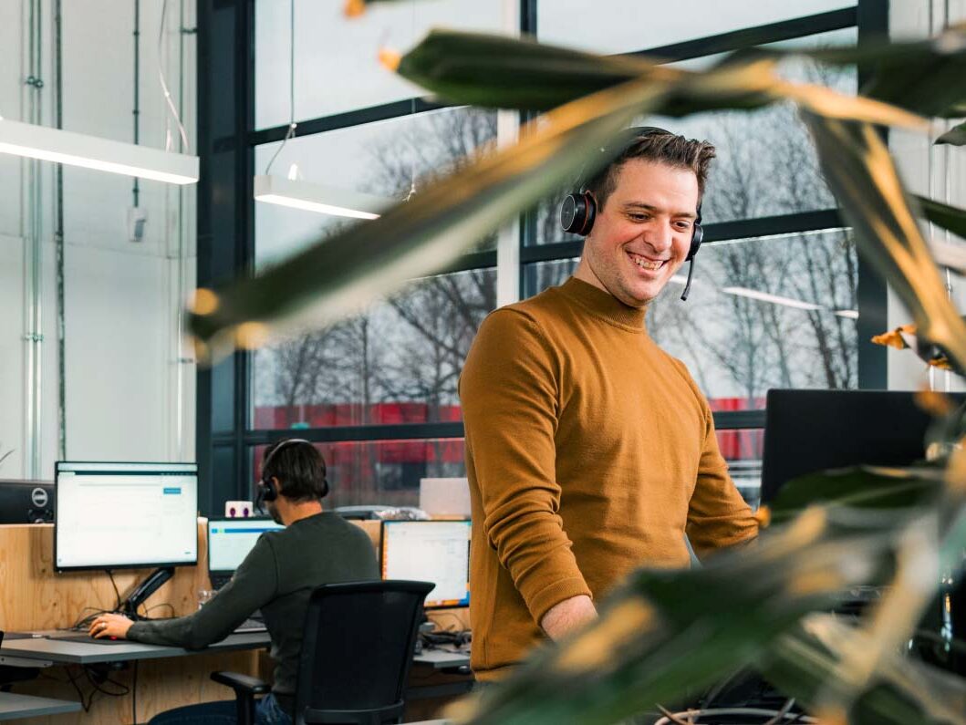 Een man met een glimlach staat aan een verstelbaar bureau in een licht en modern kantoor. Hij draagt een mosterdkleurige trui en een koptelefoon met microfoon. Achter hem zitten collega's geconcentreerd achter hun computerschermen. Het kantoor is voorzien van grote ramen waardoor natuurlijk daglicht binnenstroomt en is aangekleed met levendige kamerplanten, wat een frisse uitstraling geeft aan de werkruimte.