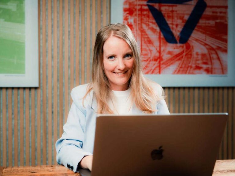 Een vriendelijk lachende vrouw met lang blond haar zit achter een laptop van Apple op een houten bureau. Ze draagt een lichtblauw jasje over een witte top. Op de achtergrond hangt een kleurrijke kunstwerk met een prominent rood en blauw design. De ruimte heeft een moderne uitstraling met een houten lambrisering op de muur