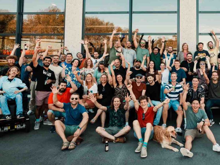 Een grote groep vrolijke mensen verzameld voor een foto, met de handen omhoog en lachende gezichten. Ze staan buiten voor een gebouw met grote ramen. Verschillende individuen maken het 'peace'-teken, sommigen hebben drankjes in hun hand. Ze dragen casual kleding in diverse stijlen en kleuren. Vooraan zit een hond ontspannen tussen de mensen. De groep straalt een gevoel van gemeenschap en plezier uit.